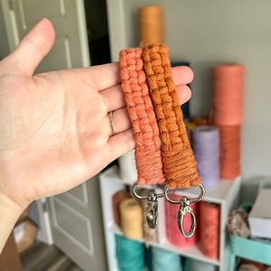 May include: Two orange macrame keychains with silver swivel clips. The keychains are made with a thick, textured cord.