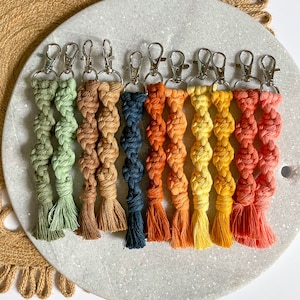 Op de afbeelding: Tien kleurrijke macramé sleutelhangers met zilveren sleutelringen. De sleutelhangers zijn gemaakt met een gedraaide touwtechniek en hebben kwastjes aan de onderkant. De kleuren zijn groen, bruin, donkerblauw, oranje, geel en rood.