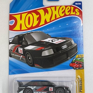Puede incluir: Un modelo a escala Hot Wheels de un Audi 90 Quattro de carreras. El coche negro presenta detalles en rojo y blanco, el número 25 y el texto "Audi Sport". El embalaje incluye el logotipo de Hot Wheels y el texto "Peak Pursuit".
