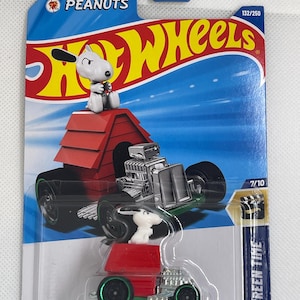 Op de afbeelding: Een Hot Wheels-gietijzeren auto met Snoopy en zijn hondenhok. De auto is zwart met een rood hondenhokdak en groene wielen. De verpakking bevat het Peanuts-logo en de tekst "HW Screen Time".
