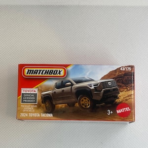 May include: A Matchbox toy car box featuring a 2024 Toyota Tacoma pickup truck. The box is red, orange, and white with the Matchbox and Toyota logos. The truck is a light brown color, and the box indicates it is an official licensed product.
