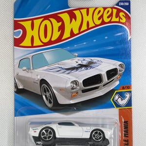 May include: A white '73 Pontiac Firebird die-cast toy car on a blue and orange Hot Wheels card. The car has blue and white graphics on the hood. The card features the Hot Wheels logo and the text "Muscle Mania".