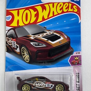 May include: A Hot Wheels die-cast toy car of a Toyota GR86 Cup in a maroon color with gold wheels. The packaging is blue and features the Hot Wheels logo and the text "GUARANTEED FOR LIFE". The car is labeled as 79/250 and 1/10 in the Compact Kings series.