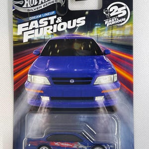 May include: A blue Hot Wheels die-cast model of a 1999 Nissan Maxima from the Fast & Furious series. The packaging features the Hot Wheels logo, the text "Dream Lineup," and "Fast & Furious." The car is displayed in a clear plastic case.