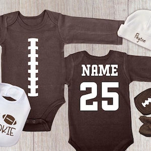 Peut inclure: Ensemble de v&ecirc;tements et accessoires pour b&eacute;b&eacute; sur le th&egrave;me du football am&eacute;ricain. Comprend un body marron avec un motif de ballon de football blanc, un bavoir avec le texte "Lil' Rookie", un bonnet blanc avec un nom et un body marron de style maillot avec "NAME 25".