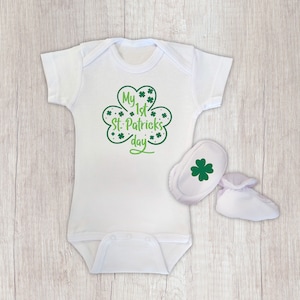 K&ouml;nnte beinhalten: Wei&szlig;er Baby-Strampler mit einem gr&uuml;nen Kleeblatt-Design, auf dem "My 1st St. Patrick's Day" steht. Der Strampler wird mit wei&szlig;en Schuhen mit einem gr&uuml;nen Kleeblatt-Design geliefert.