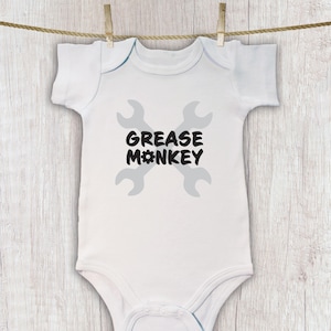 May include: White baby onesie with the text "GREASE MONKEY" in black, with a gray wrench design. The onesie is hanging from a clothesline with wooden clothespins against a light wood background.
