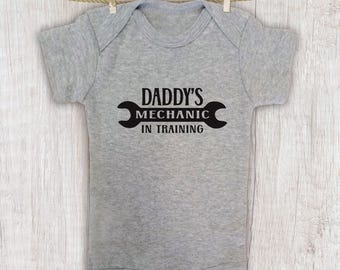 May include: A gray baby onesie with the text "DADDY'S MECHANIC IN TRAINING" in black. The onesie is hanging from a clothesline with wooden clothespins. The design includes a wrench graphic.