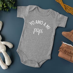 May include: A gray bodysuit with the text "YO AMO A MI papi" in white. The bodysuit is on a blue background with a stuffed bunny, brown booties, and a yellow ribbon.