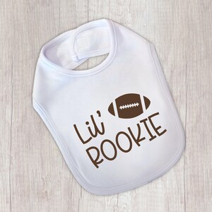 Peut inclure: Bavoir de b&eacute;b&eacute; blanc avec un ballon de football am&eacute;ricain marron et l'inscription "Lil' ROOKIE" en marron. Le bavoir est de forme arrondie et poss&egrave;de une fermeture velcro au cou.