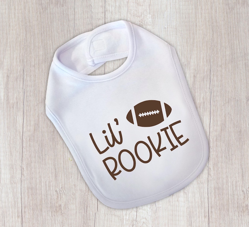 Peut inclure: Bavoir de b&eacute;b&eacute; blanc avec un ballon de football am&eacute;ricain marron et l'inscription "Lil' ROOKIE" en marron. Le bavoir est de forme arrondie et poss&egrave;de une fermeture velcro au cou.
