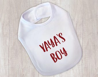 May include: White baby bib with the text "YAYA'S BOY" in a bold, burgundy font. The bib has a rounded shape with a Velcro closure at the neck. The bib is made of a soft material, perfect for catching spills and drool.