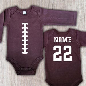 Peut inclure: Deux bodies de b&eacute;b&eacute; &agrave; manches longues de couleur bordeaux. L'un pr&eacute;sente un motif de ballon de football blanc, l'autre porte le mot "NAME" au-dessus du num&eacute;ro "22" en blanc. Pour nourrissons.