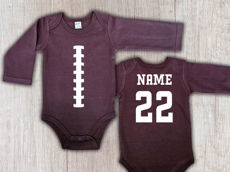Peut inclure: Deux bodies de b&eacute;b&eacute; &agrave; manches longues de couleur bordeaux. L'un pr&eacute;sente un motif de ballon de football blanc, l'autre porte le mot "NAME" au-dessus du num&eacute;ro "22" en blanc. Pour nourrissons.
