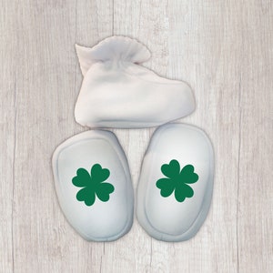 K&ouml;nnte beinhalten: Drei wei&szlig;e Babyschuhe mit gr&uuml;nen Kleeblatt-Designs. Die Schuhe liegen auf einem hellen Holzhintergrund.