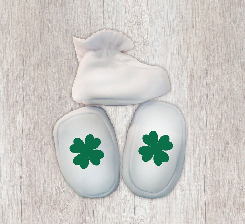 K&ouml;nnte beinhalten: Drei wei&szlig;e Babyschuhe mit gr&uuml;nen Kleeblatt-Designs. Die Schuhe liegen auf einem hellen Holzhintergrund.