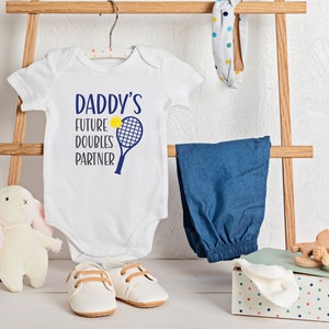 May include: White baby onesie with the text "Daddy's Future Doubles Partner" and a blue tennis racket with a yellow tennis ball. The onesie is hanging on a wooden rack with a blue pair of pants, a white headband with colorful polka dots, a white stuffed bunny, and a wooden toy.
