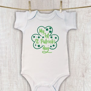 K&ouml;nnte beinhalten: Wei&szlig;er Baby-Strampler mit einem gr&uuml;nen Kleeblatt-Design und dem Text "My 1st St. Patrick's Day".