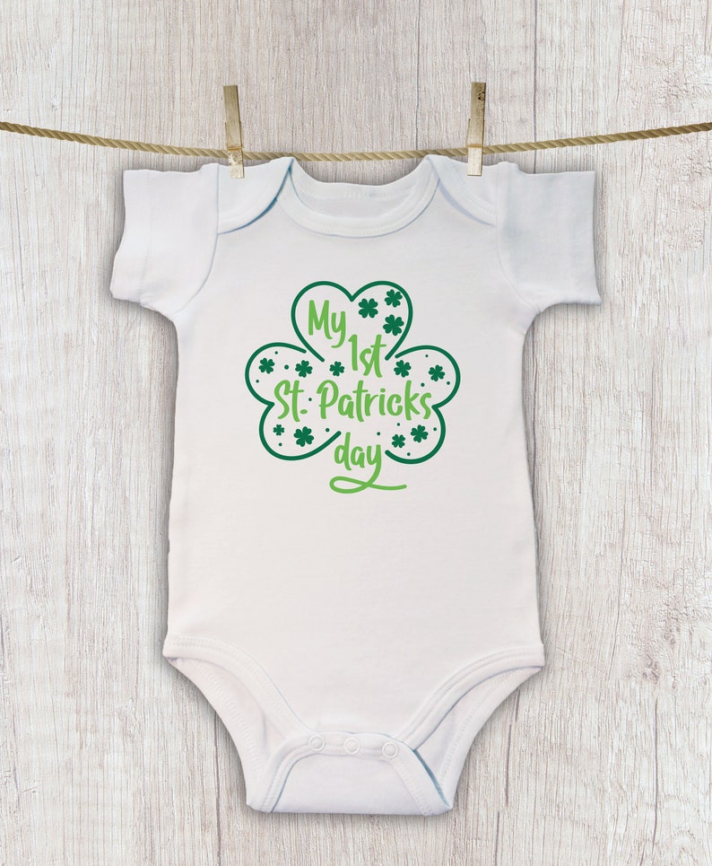 K&ouml;nnte beinhalten: Wei&szlig;er Baby-Strampler mit einem gr&uuml;nen Kleeblatt-Design und dem Text "My 1st St. Patrick's Day".