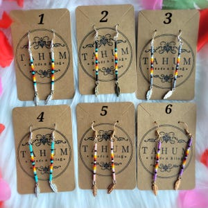 May include: Six pairs of beaded earrings with feather charms are displayed on brown cards. Each pair features a different color scheme, including turquoise, black, and purple beads. The cards have the text "TAHUM Beads & Bling."