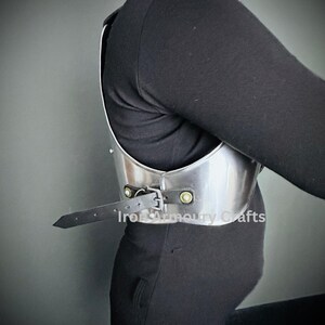 May include: A silver metal chest plate with a black leather strap and metal buckles. The chest plate is worn by a person and the strap is fastened at the side. The text "Iron Armoury Crafts" is visible on the chest plate.