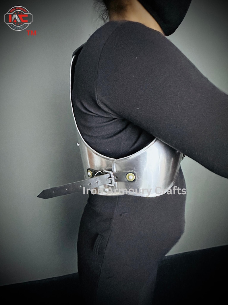 May include: A silver metal chest plate with a black leather strap and metal buckles. The chest plate is worn by a person and the strap is fastened at the side. The text "Iron Armoury Crafts" is visible on the chest plate.