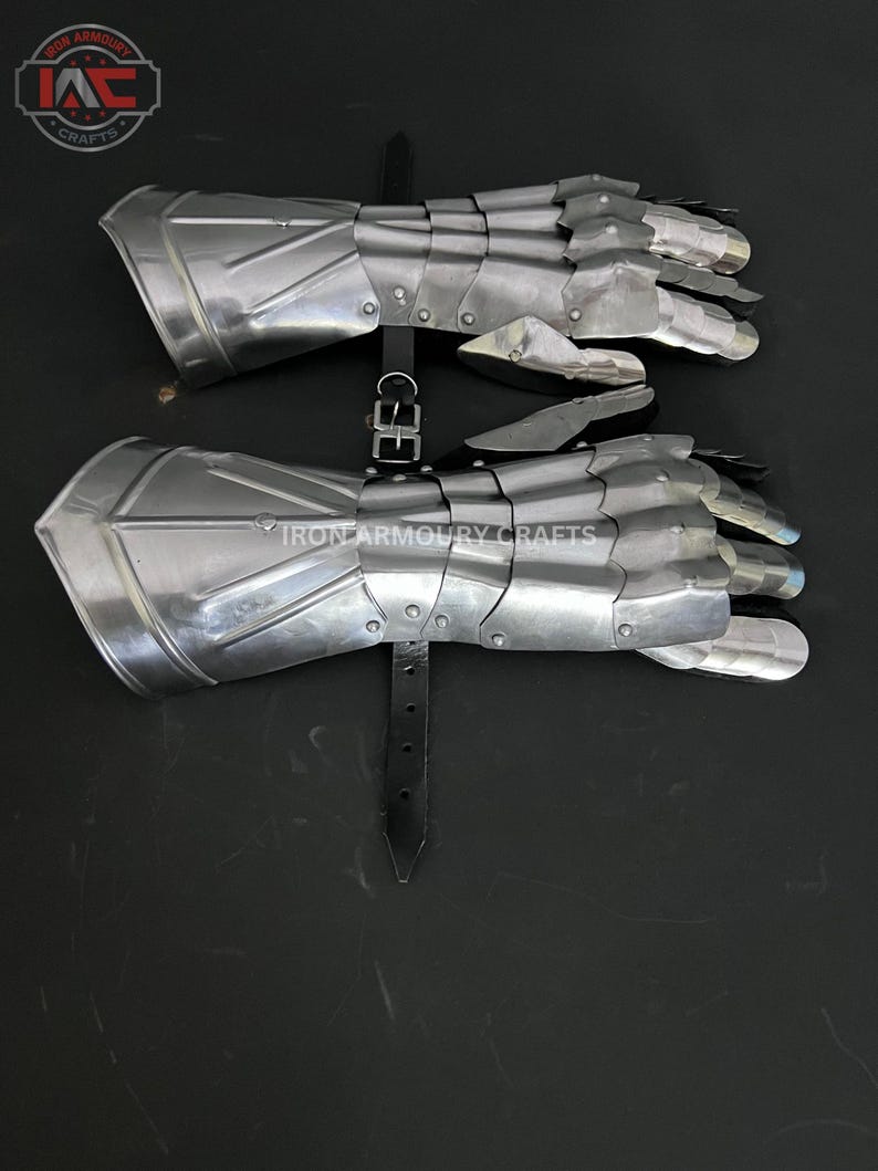 Op de afbeelding: Een paar gearticuleerde metalen handschoenen, waarschijnlijk voor historische re-enactment. De zilverkleurige handschoenen zijn gemaakt van overlappende platen, met leren riemen en gespen om ze vast te zetten. Het Iron Armoury Crafts logo is zichtbaar.