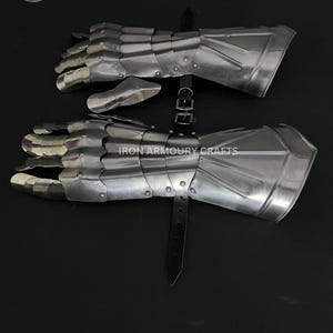 Op de afbeelding: Een paar zilverkleurige, gearticuleerde metalen handschoenen, mogelijk voor historische doeleinden. De handschoenen hebben individuele vingerplaten en een manchet. De tekst "IRON ARMOURY CRAFTS" is zichtbaar. Een zwarte leren riem zet de handschoenen vast.