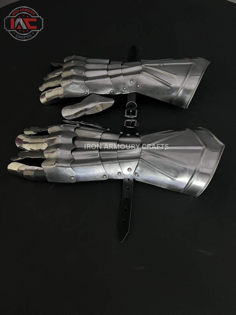 Op de afbeelding: Een paar zilverkleurige, gearticuleerde metalen handschoenen, mogelijk voor historische doeleinden. De handschoenen hebben individuele vingerplaten en een manchet. De tekst "IRON ARMOURY CRAFTS" is zichtbaar. Een zwarte leren riem zet de handschoenen vast.