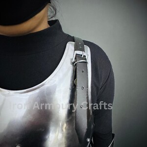 May include: A silver metal chest plate with a black leather strap. The chest plate is worn by a person wearing a black shirt. The text "Iron Armoury Crafts" is visible on the chest plate.