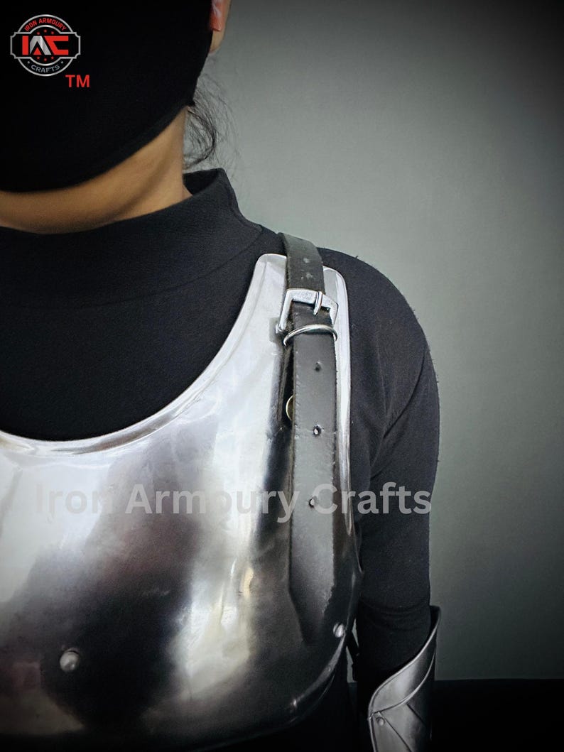 May include: A silver metal chest plate with a black leather strap. The chest plate is worn by a person wearing a black shirt. The text "Iron Armoury Crafts" is visible on the chest plate.
