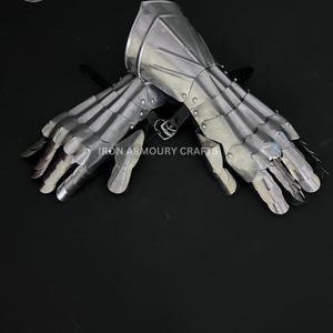 Op de afbeelding: Een paar zilverkleurige metalen handschoenen, met beweeglijke vingersegmenten en beschermende platen. De handschoenen zijn ontworpen in een middeleeuwse stijl, met de tekst "IRON ARMOURY CRAFTS" zichtbaar. Ze liggen op een donkere ondergrond.