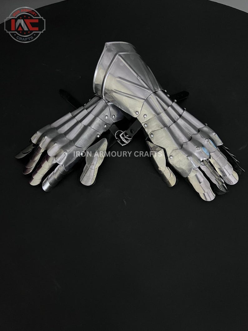 Op de afbeelding: Een paar zilverkleurige metalen handschoenen, met beweeglijke vingersegmenten en beschermende platen. De handschoenen zijn ontworpen in een middeleeuwse stijl, met de tekst "IRON ARMOURY CRAFTS" zichtbaar. Ze liggen op een donkere ondergrond.