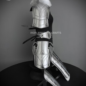 May include: Silver plated armor set for the legs and arms. The set includes a pair of shin guards, a pair of knee pads, a pair of elbow pads, and a pair of shoulder pads. The armor is made of metal and has a polished finish. The armor is displayed on a black stand. "IRON ARMOURY CRAFTS" is printed on the stand.