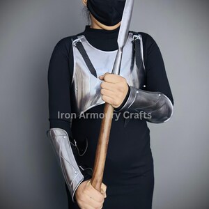 May include: A person wearing a black shirt and a face mask is holding a silver spear and wearing silver armor on their arms and chest. The armor has straps and rivets. The spear has a wooden shaft and a silver blade. The person is standing in front of a gray wall. The text "Iron Armoury Crafts" is visible on the image.