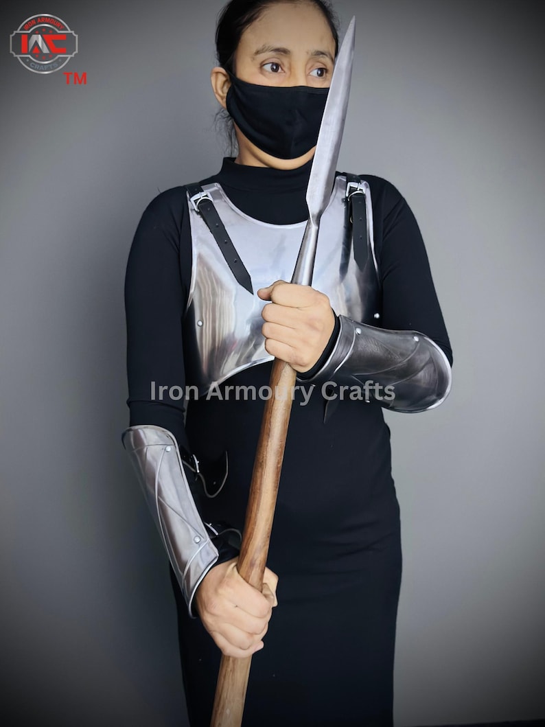 May include: A person wearing a black shirt and a face mask is holding a silver spear and wearing silver armor on their arms and chest. The armor has straps and rivets. The spear has a wooden shaft and a silver blade. The person is standing in front of a gray wall. The text "Iron Armoury Crafts" is visible on the image.
