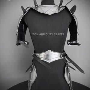 May include: Silver and black armor for the torso, shoulders, and hips. The armor is made of metal plates and leather straps. The armor is displayed on a black mannequin. The text "IRON ARMOURY CRAFTS" is printed on the mannequin.