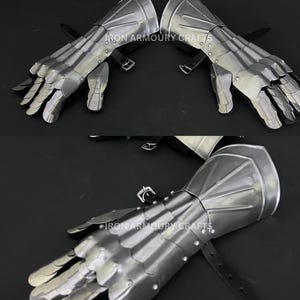 Op de afbeelding: Een paar gepolijste stalen handschoenen met beweeglijke vingers en handbescherming. De handschoenen hebben een metallic glans en zijn vastgezet met leren riemen en gespen. De tekst "IRON ARMOURY CRAFTS" is zichtbaar.