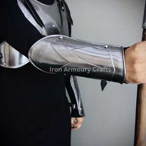 May include: A silver metal arm guard with a pointed edge and a strap. The arm guard is worn on the left arm of a person wearing a black shirt and a silver metal chest piece. The text "Iron Armoury Crafts" is visible on the arm guard.