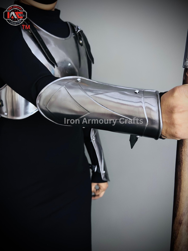 May include: A silver metal arm guard with a pointed edge and a strap. The arm guard is worn on the left arm of a person wearing a black shirt and a silver metal chest piece. The text "Iron Armoury Crafts" is visible on the arm guard.