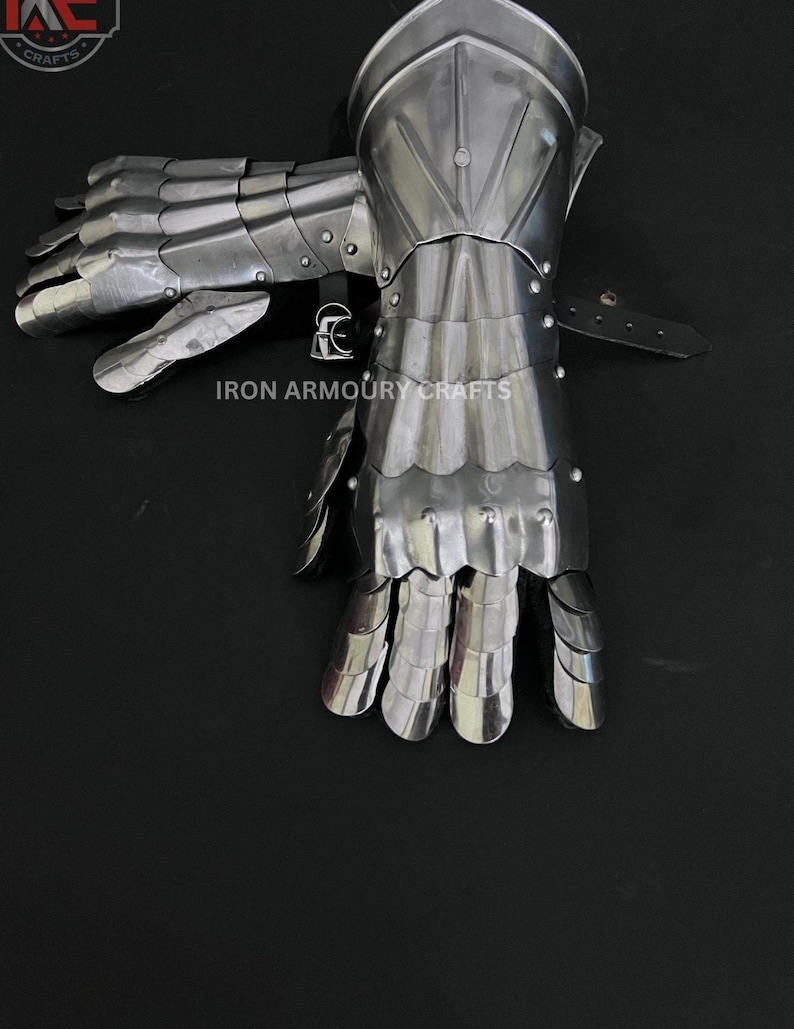 Op de afbeelding: Een paar gearticuleerde stalen handschoenen, waarschijnlijk voor historische re-enactment. De handschoenen zijn zilverkleurig, met overlappende platen en klinknagels. Een leren riem is zichtbaar. De afbeelding is tegen een donkere achtergrond.