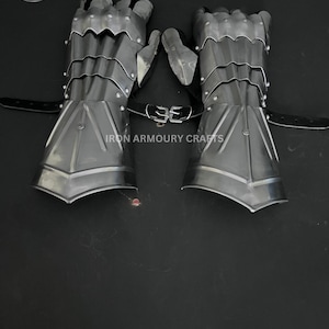 Op de afbeelding: Een paar zilverkleurige metalen handschoenen met beweeglijke vingers en polsbescherming. De handschoenen zijn verbonden door een zwarte leren riem met een gesp. De tekst "IRON ARMOURY CRAFTS" staat op de afbeelding.