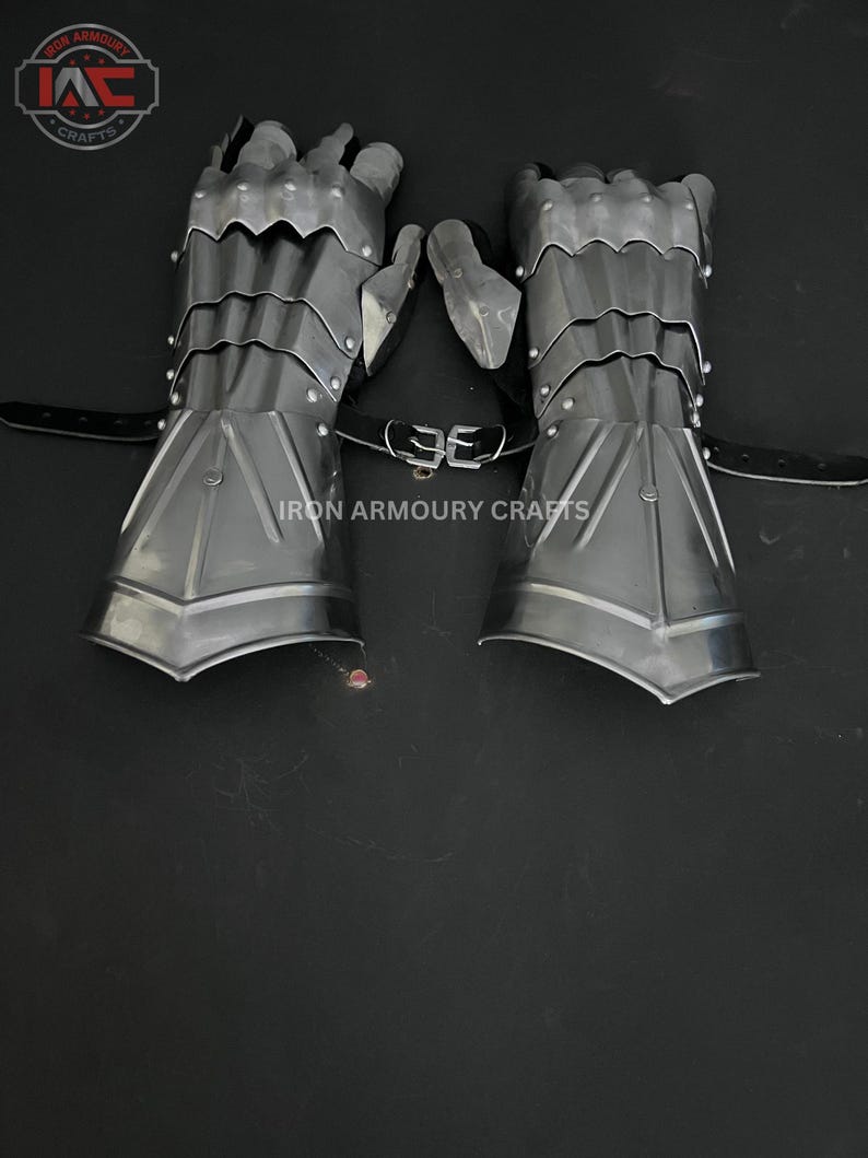 Op de afbeelding: Een paar zilverkleurige metalen handschoenen met beweeglijke vingers en polsbescherming. De handschoenen zijn verbonden door een zwarte leren riem met een gesp. De tekst "IRON ARMOURY CRAFTS" staat op de afbeelding.