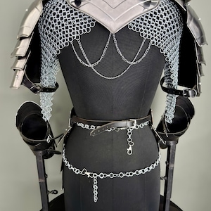 May include: A medieval-style suit of armor displayed on a black mannequin. The armor features a silver breastplate, shoulder guards, and chainmail. A black leather belt and silver chain belt are also included. The armor is designed for protection and display.
