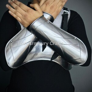 May include: A pair of silver metal arm guards with a decorative design. The guards are worn on the forearms and extend to the wrists.