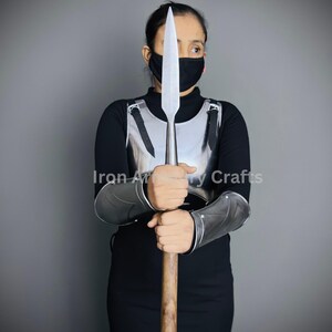 May include: A person wearing a black shirt and a black face mask is holding a silver spear with a wooden shaft. The person is also wearing a silver chest plate and silver arm guards. The text "Iron Armory Crafts" is visible in the image.