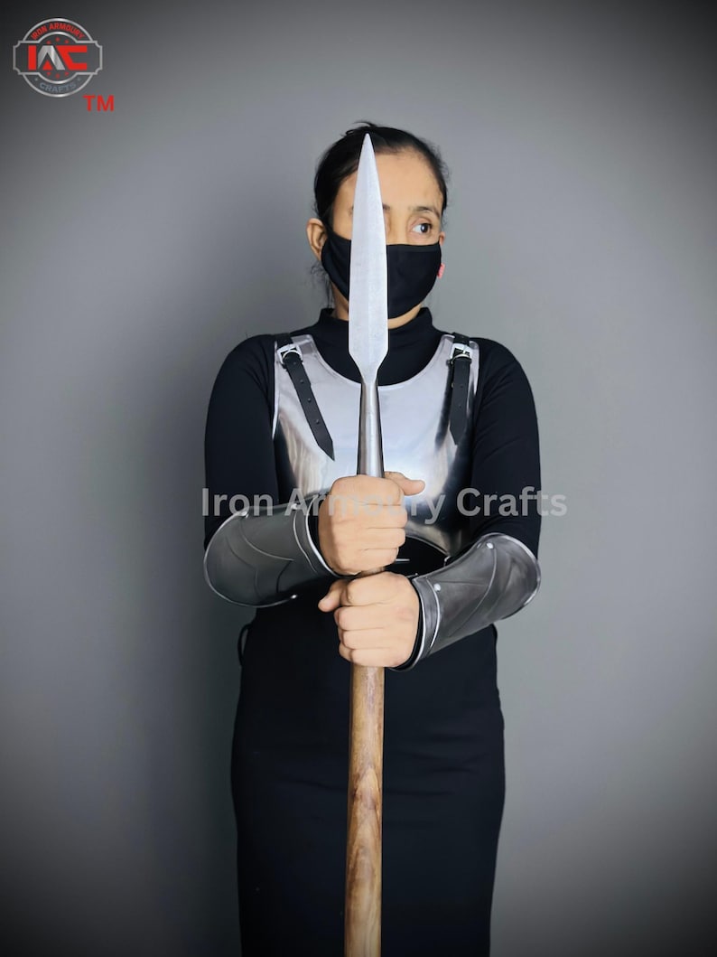 May include: A person wearing a black shirt and a black face mask is holding a silver spear with a wooden shaft. The person is also wearing a silver chest plate and silver arm guards. The text "Iron Armory Crafts" is visible in the image.