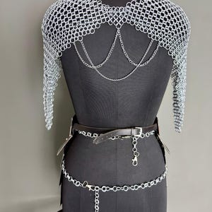 May include: A silver chainmail shoulder piece with long sleeves, displayed on a black mannequin. The chainmail is connected with silver chains that hang down the back. A black leather belt and silver chain belt are also featured.
