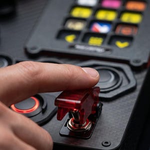 A black and red control panel with a carbon fiber finish. The panel has a variety of buttons, knobs, and switches. The text "RACEBOX" is printed on the panel. The text "REAL CARBON FIBER" is printed on a yellow sticker.