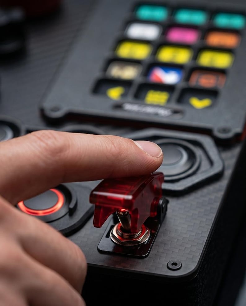 A black and red control panel with a carbon fiber finish. The panel has a variety of buttons, knobs, and switches. The text "RACEBOX" is printed on the panel. The text "REAL CARBON FIBER" is printed on a yellow sticker.